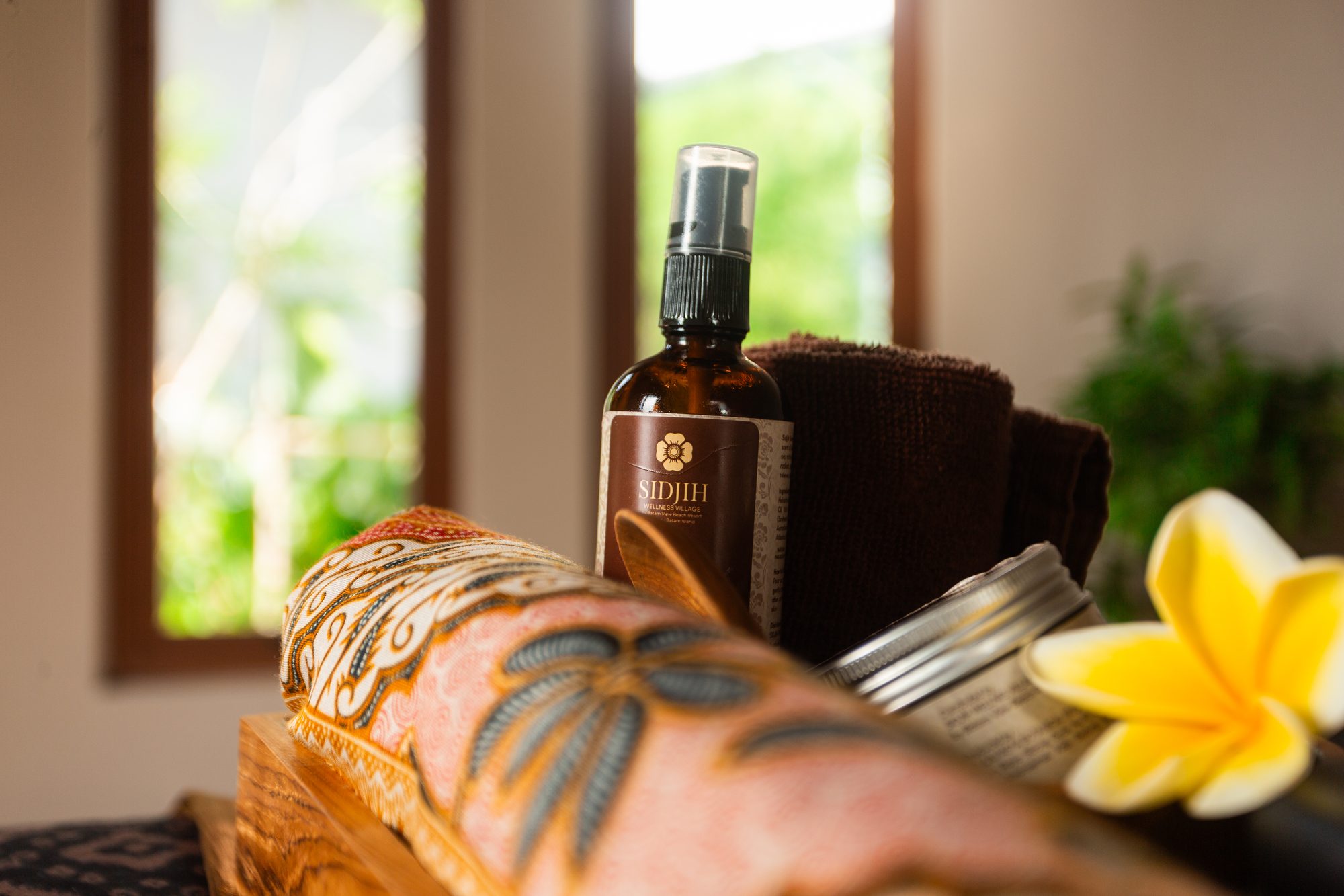 A bottle of Sidjih massage oil beside folded towels and a frangipani blossom